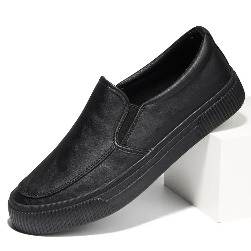 A black men’s slip-on casual shoe with a hybrid rubber sole, displayed at an angle with the heel resting on a white block. The shoe features a sleek, plain design, visible stitching along the sides, and a comfortable leather insole.