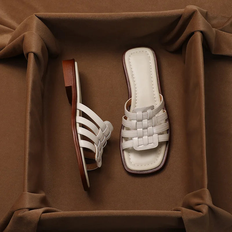 A pair of white woven Italian leather slide sandals with a square-toe design, displayed on a padded brown fabric surface—one sandal laying flat and the other resting on its side.