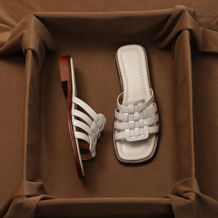 A pair of white woven Italian leather slide sandals with a square-toe design, displayed on a padded brown fabric surface—one sandal laying flat and the other resting on its side.