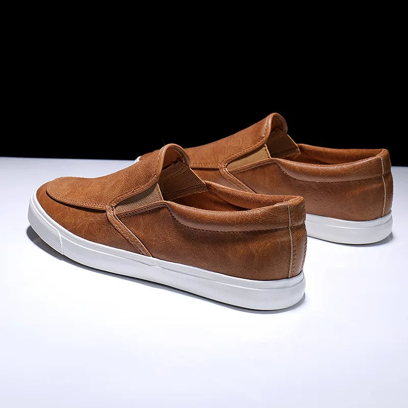 A pair of mens slip-on brown casual shoes with a leather insole and white rubber-leather sole, displayed on a white surface against a black background.