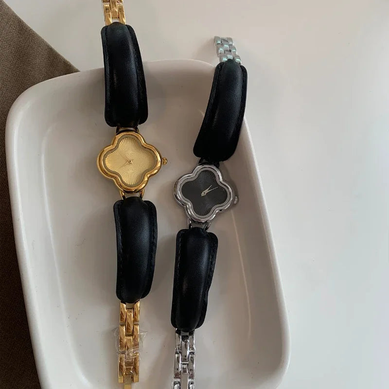 Two clover dial watches—one luxury gold-plated with a gold band, the other silver with a black face and silver band—are displayed on a white tray with black cushions under their bands.