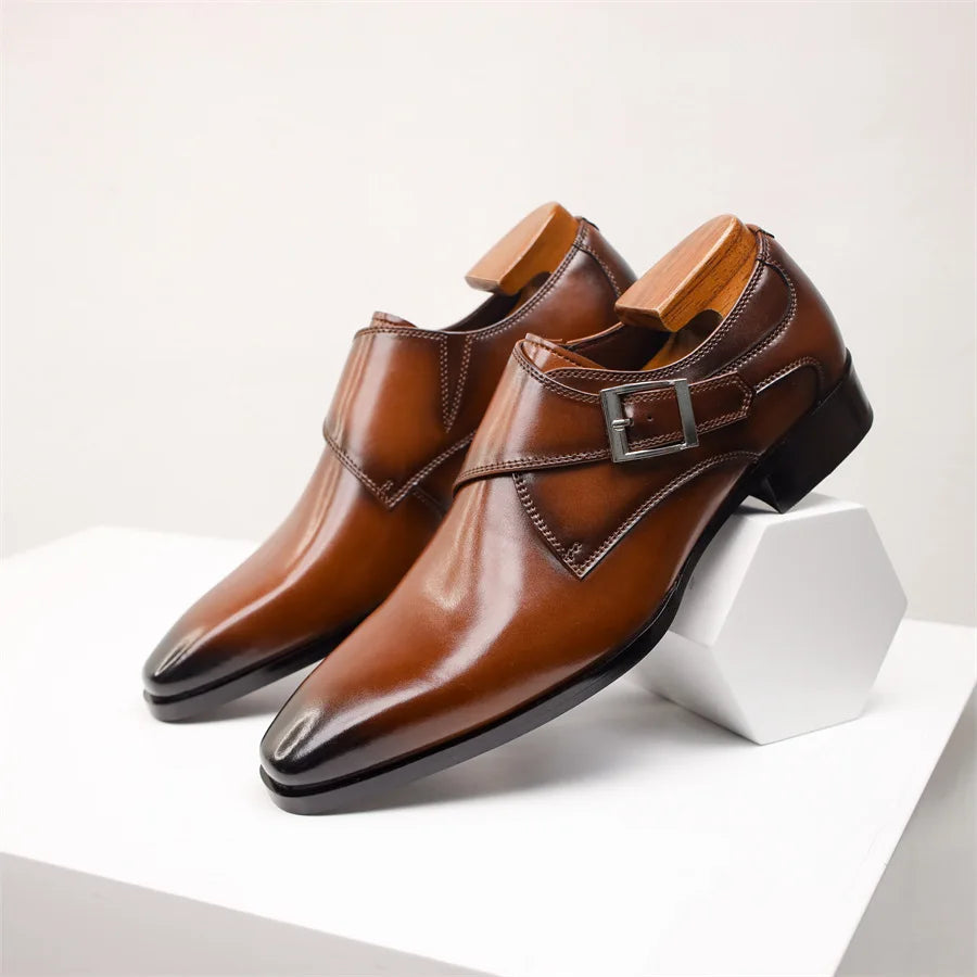 A pair of polished brown Italian leather monk strap shoes with buckles, displayed on a white geometric stand against a plain background. Shoe trees are inserted to maintain their shape.