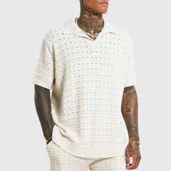 A person with tattoos on their neck and arms wears a cream-colored, short-sleeve summer shirt with a collar and matching knit pants. The shirt features a breathable knit design with a textured, perforated pattern. The background is plain light gray.