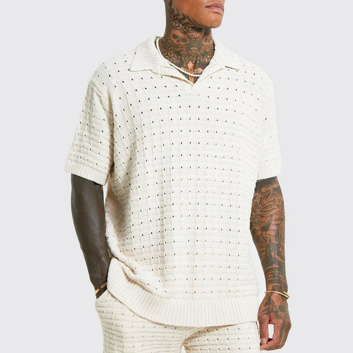 A person with tattoos on their neck and arms wears a cream-colored, short-sleeve summer shirt with a collar and matching knit pants. The shirt features a breathable knit design with a textured, perforated pattern. The background is plain light gray.