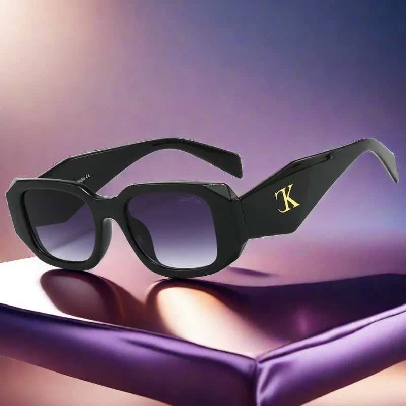 Black square sunglasses with thick frames and dark lenses rest on a shiny surface. The left arm displays a prominent gold K logo. These oversized acetate sunglasses offer UV400 protection against harmful rays, set against a soft purple and beige gradient background.