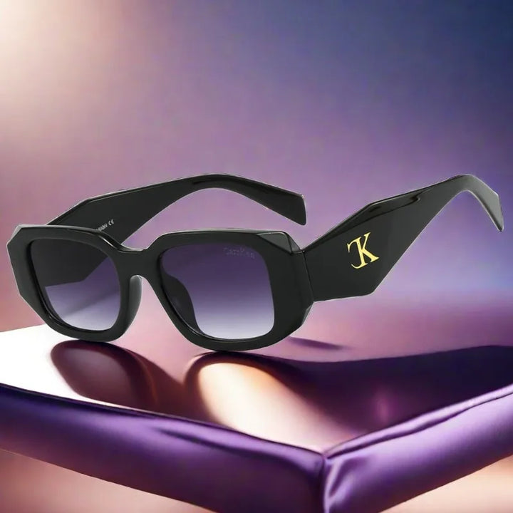 Black square sunglasses with thick frames and dark lenses rest on a shiny surface. The left arm displays a prominent gold K logo. These oversized acetate sunglasses offer UV400 protection against harmful rays, set against a soft purple and beige gradient background.