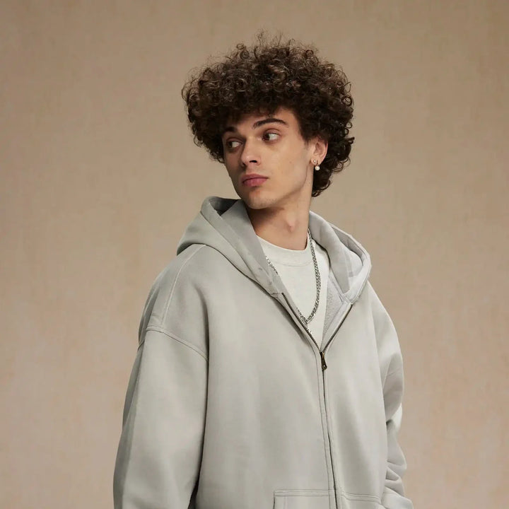 A young person with curly brown hair wearing a relaxed fit gray hoodie and a chain necklace looks to the side against a plain beige background.