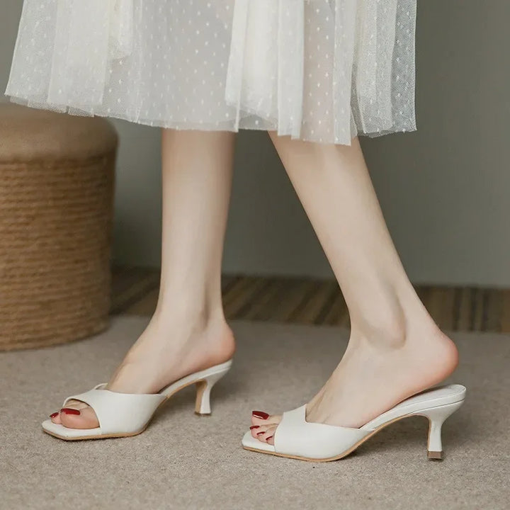 A person wearing a sheer white polka dot dress and white open-toe heeled sandals with low heel and ergonomic arch support stands indoors on a carpeted floor.