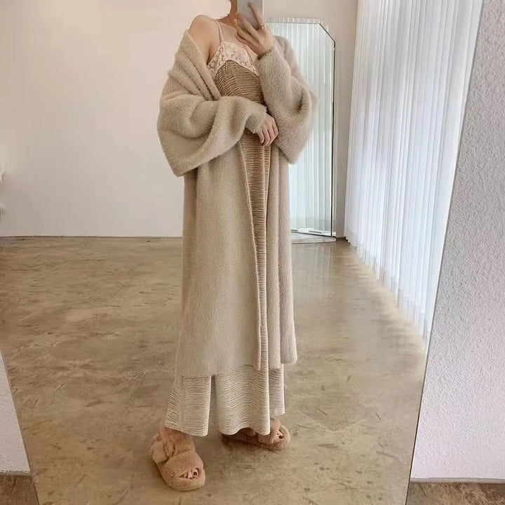 A person stands in front of a mirror wearing a long, beige, cozy cardigan with a relaxed drape fit over a light dress, and fluffy beige slippers. The room has minimal decor with white curtains and a polished floor.