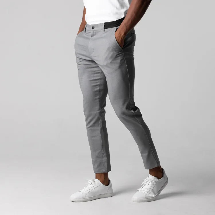 A person wearing light gray slim-fit, ankle length pants with 4 way stretch, a white t-shirt, and white sneakers stands with hands in pockets against a plain gray background. The image shows only the lower half of the body.