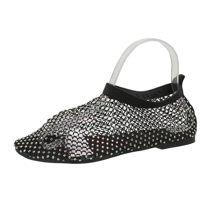 A black flat shoe featuring dazzling rhinestone accents, a mesh overlay upper design, transparent heel strap, and a cushioned footbed for added comfort.