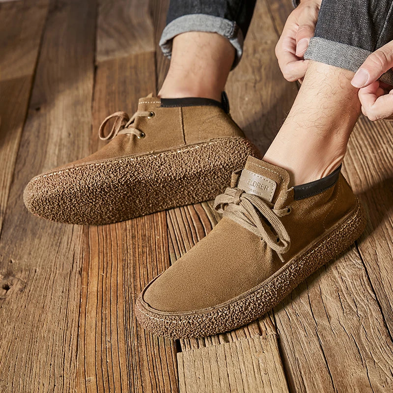 A person wearing rolled-up jeans is putting on light brown Men's Chaussure Homme Shoes made of cow suede leather with textured rubber soles, sitting on a wooden floor.