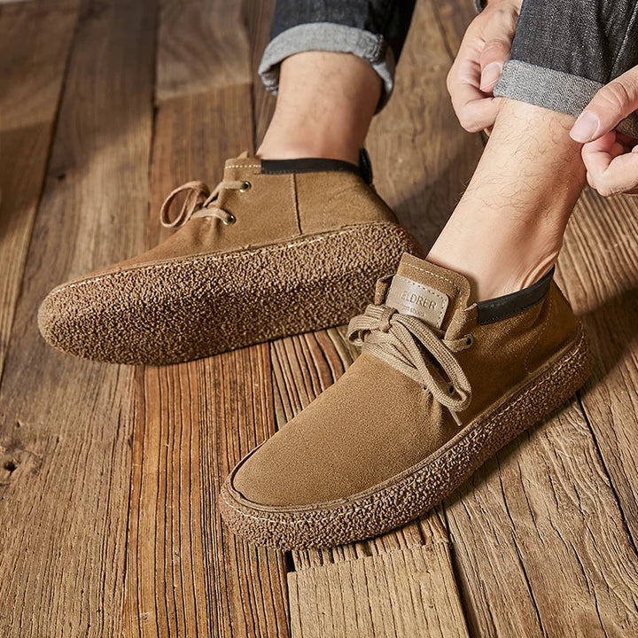 A person wearing rolled-up jeans is putting on light brown Men's Chaussure Homme Shoes made of cow suede leather with textured rubber soles, sitting on a wooden floor.