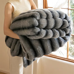 A person in light clothing holds a large, thick, luxurious throw with an elegant wave pattern in a sunlit room with a window and natural light in the background.