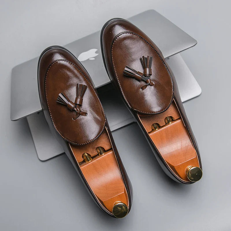 A pair of brown leather tassel loafers with a cushioned insole and wooden shoe trees rests on top of a closed silver laptop with an Apple logo, on a gray surface.