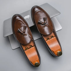 A pair of brown leather tassel loafers with a cushioned insole and wooden shoe trees rests on top of a closed silver laptop with an Apple logo, on a gray surface.