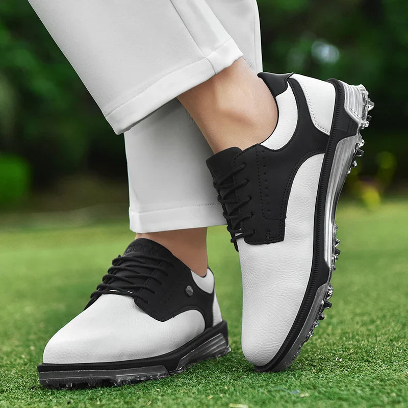 A person wearing white pants and stylish black and white golf shoes with a high-traction outsole stands on green grass, one foot slightly crossed over the other. The shoes feature black soles and black laces.