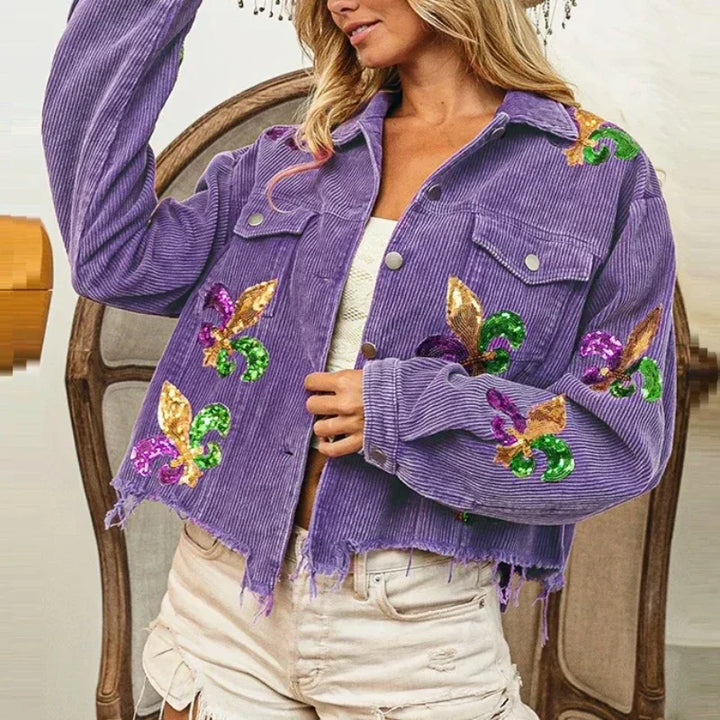 A woman in a frayed purple corduroy jacket with sequin floral patches and light distressed shorts sits in a chair. The cropped relaxed fit jacket stands out against the neutral background with wooden details.