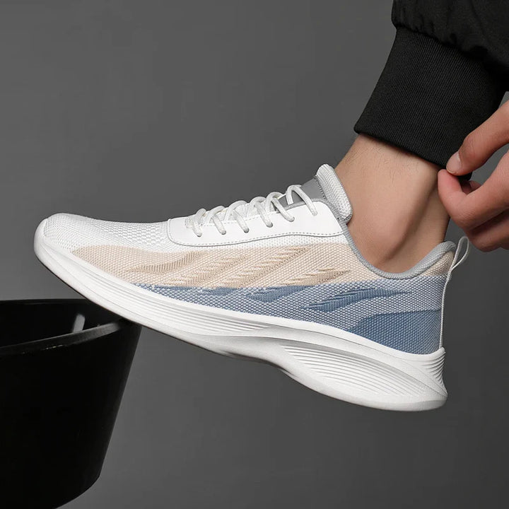 A person wearing a stylish sneaker with white, beige, and blue accents featuring breathable mesh, shown in profile against a gray background. The person is dressed in black pants and is raising their foot, highlighting the shoe’s design.