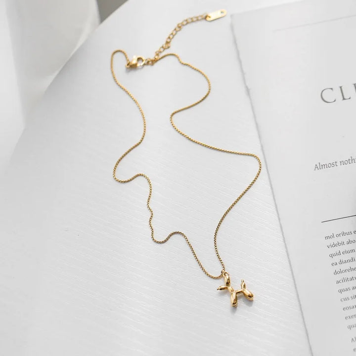 A delicate gold balloon dog necklace with a playful jewelry vibe rests on a white textured surface next to an open book or magazine.