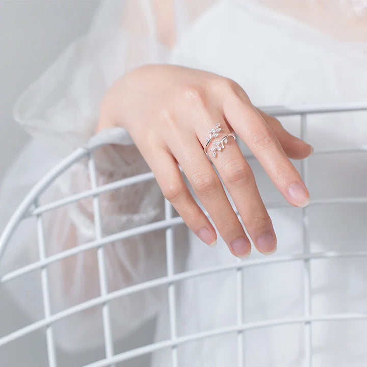 A delicate hand wearing the Isadora Floral Ring, a piece of fine jewelry with leaf designs, rests on a white wire chair. The person is dressed in a sheer white, long-sleeved garment.