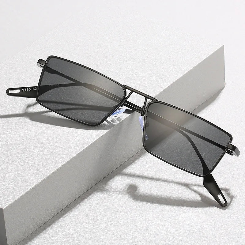 A pair of Avalon Sunglasses with black rectangular lenses and dark shades rests on a white textured surface, the arms open and slightly elevated.