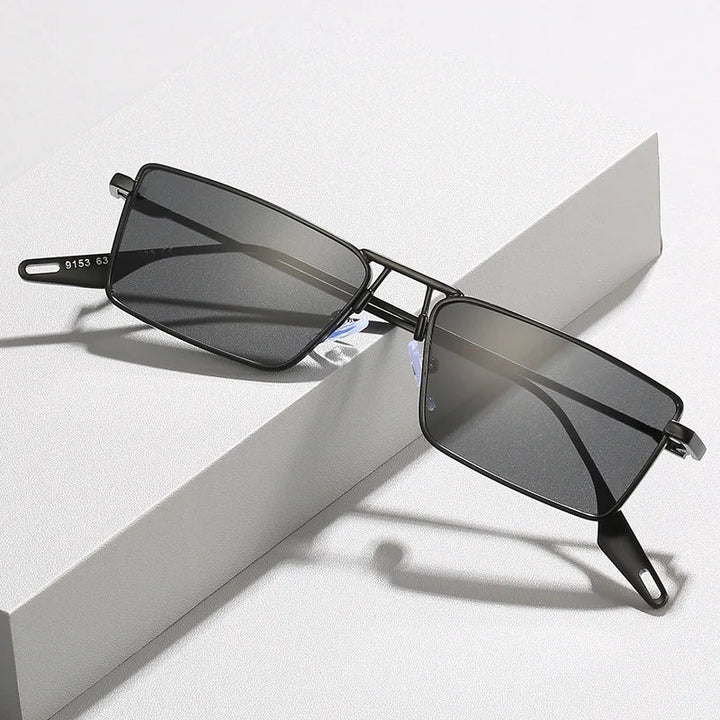 A pair of Avalon Sunglasses with black rectangular lenses and dark shades rests on a white textured surface, the arms open and slightly elevated.