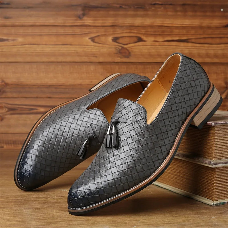 A pair of black real leather shoes with a basketweave pattern and tassels on the front, displayed on wooden surfaces with one shoe propped on books. The background features rich wood paneling, highlighting this luxury footwear.