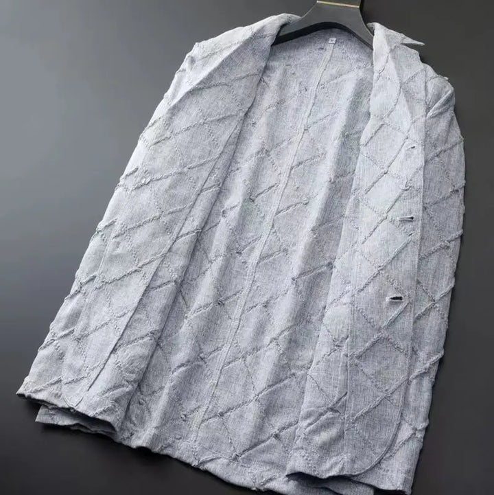 A light gray textured blazer with a quilted diamond pattern hangs on a black hanger against a dark background. The unbuttoned blazer has a slightly wrinkled, casual look, perfect for pairing with relaxed fit trousers.