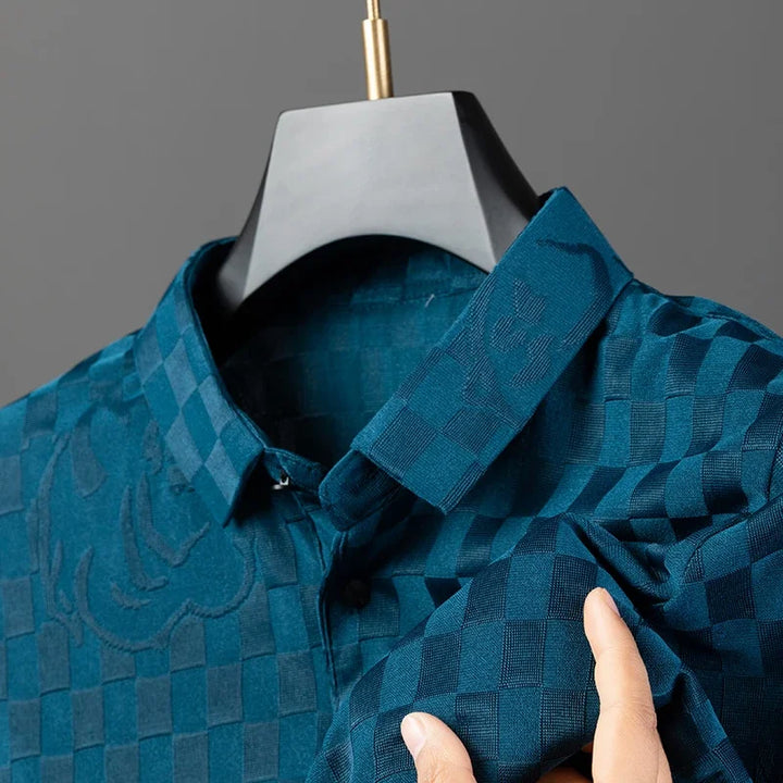 A hand holds the sleeve of a blue, tailored fit button-up shirt with subtle textured patterns and a checkerboard pattern, displayed on a black hanger against a gray background.