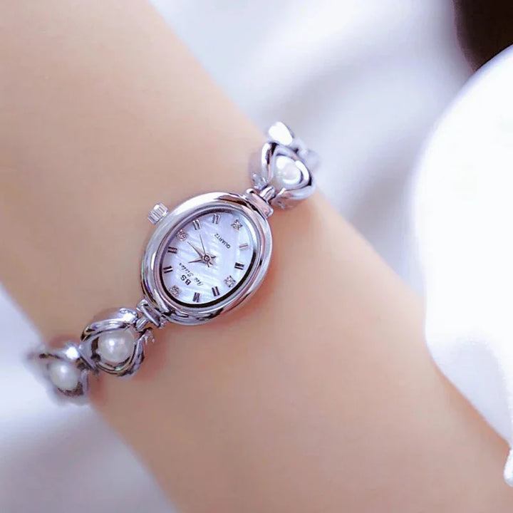 A close-up of a silver wristwatch with pearl accents on the bracelet, worn on a persons wrist. The oval watch face features black Roman numerals and silver hands, set against a soft, blurred white background.