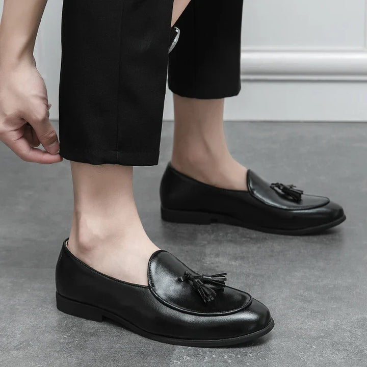 A person wearing black dress pants is putting on black leather loafers with tassel details and a cushioned insole, standing on a gray floor near a white wall.