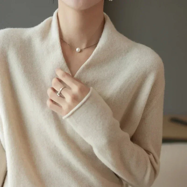 A person wearing a soft, light beige, cashmere long-sleeve sweater holds the collar with one hand. They wear a simple pearl necklace and a silver ring. The background is neutral and out of focus.