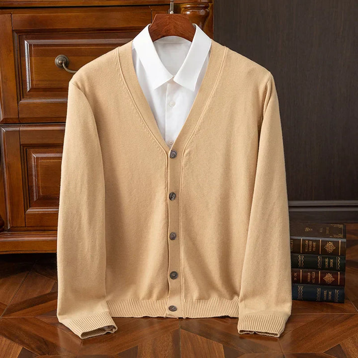 A beige cashmere wool blend button-up cardigan with a relaxed fit and a white collared shirt underneath, hanging on a wooden hanger in front of wooden drawers and a stack of books on a wooden floor.