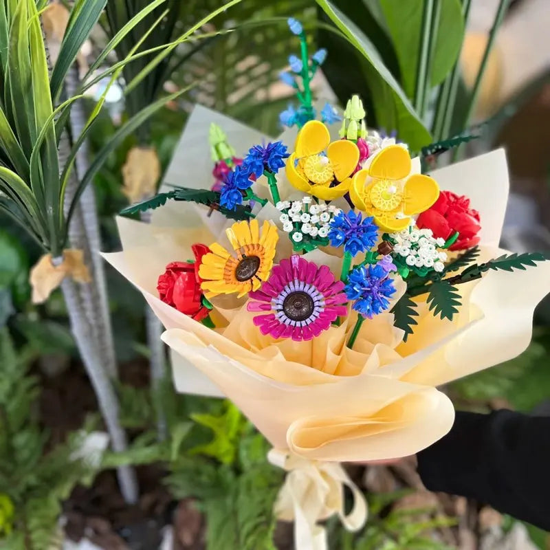 A colorful Wildflower Bouquet made from LEGO building blocks, featuring yellow, purple, blue, and red flowers with green leaves, wrapped in beige paper and tied with a ribbon, held in front of green plants.