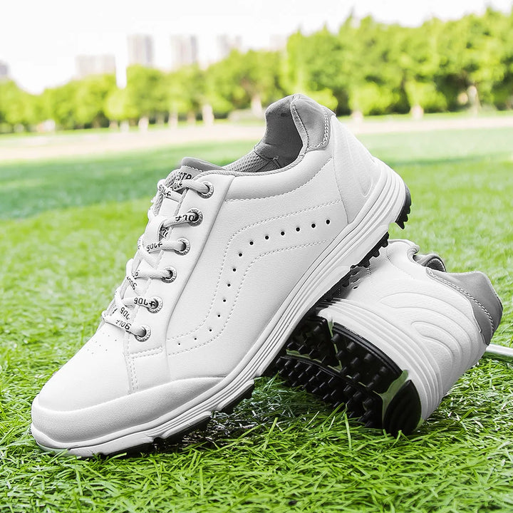 A pair of white waterproof golf shoes with gray accents are displayed on green grass, one upright and the other resting on its side. Designed for performance, this golf footwear features advanced cleat technology for superior grip. Trees and buildings blur in the background.