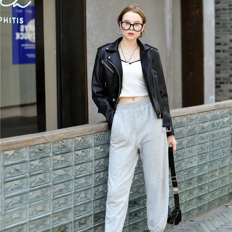 A woman wearing large glasses, a black leather jacket, a white crop top, and gray sweatpants with zipper pockets stands outdoors by a glass brick wall, holding a black bag and looking at the camera.