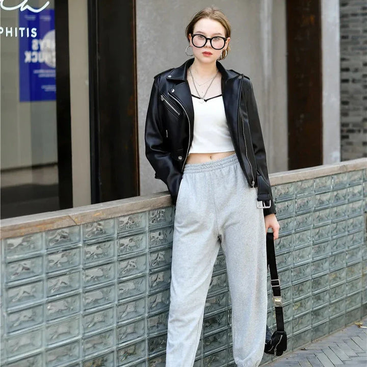 A woman wearing large glasses, a black leather jacket, a white crop top, and gray sweatpants with zipper pockets stands outdoors by a glass brick wall, holding a black bag and looking at the camera.
