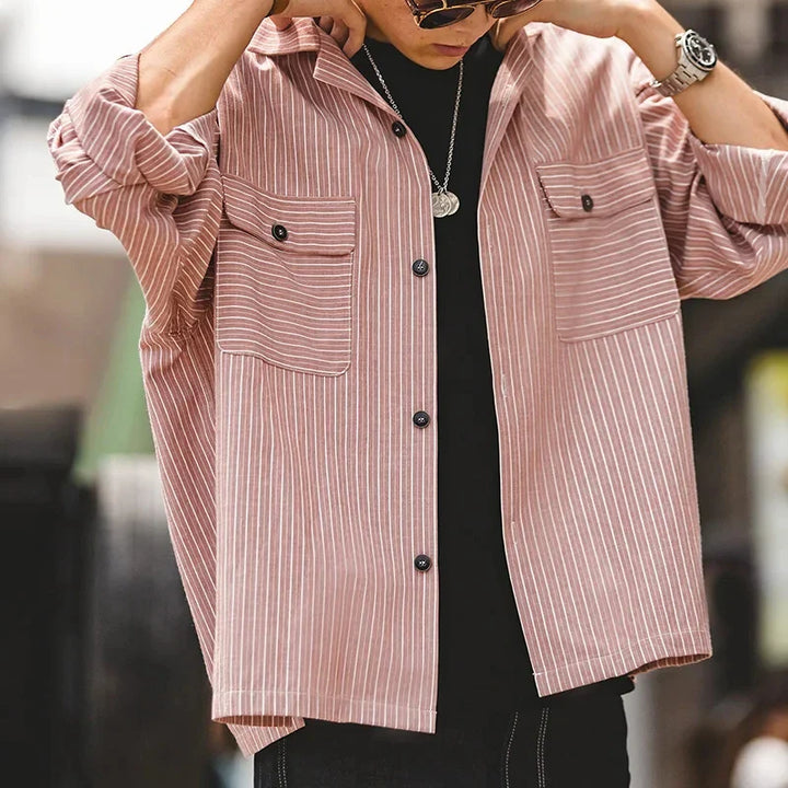 A person wearing a loose, light pink and white pinstripe Cuban collar shirt over a black t-shirt, accessorized with a silver necklace and watch, with sleeves rolled up and hands adjusting the collar.