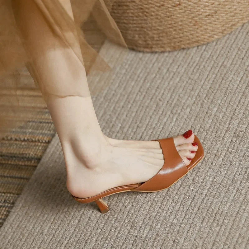 A person wearing a brown genuine leather open-toe heeled sandal with red nail polish on their toenails, standing on a textured beige carpet.