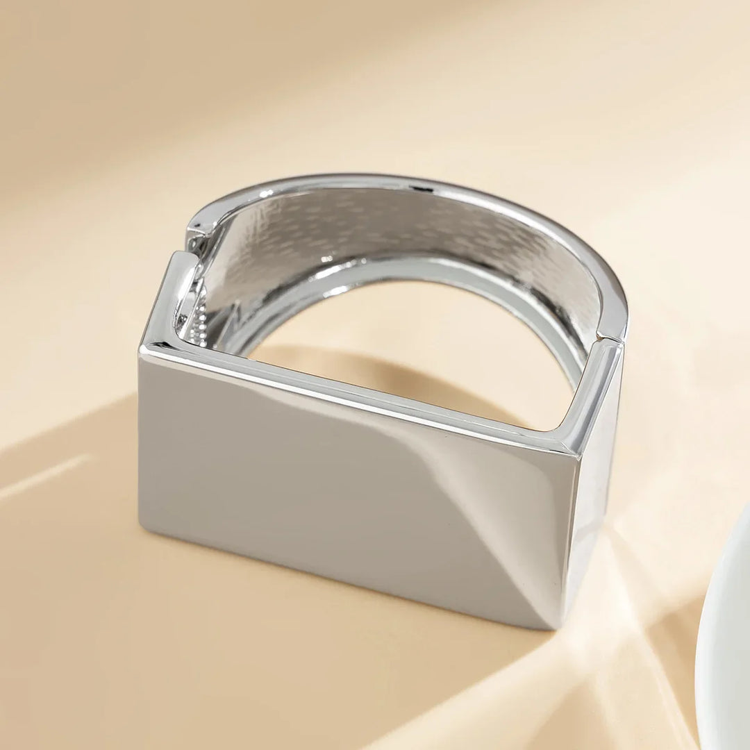 A modern, shiny silver bracelet with a geometric, rectangular design rests on a light beige surface. This unisex jewelry piece features a smooth, polished finish and an open, angular shape.