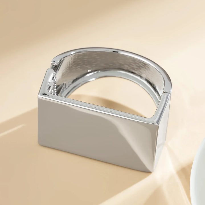 A modern, shiny silver bracelet with a geometric, rectangular design rests on a light beige surface. This unisex jewelry piece features a smooth, polished finish and an open, angular shape.