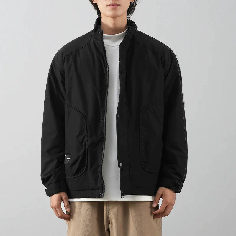 A person wearing a minimalist jacket with a black exterior over a white t-shirt and beige pants stands against a plain light gray background. The individuals face is partially out of frame.