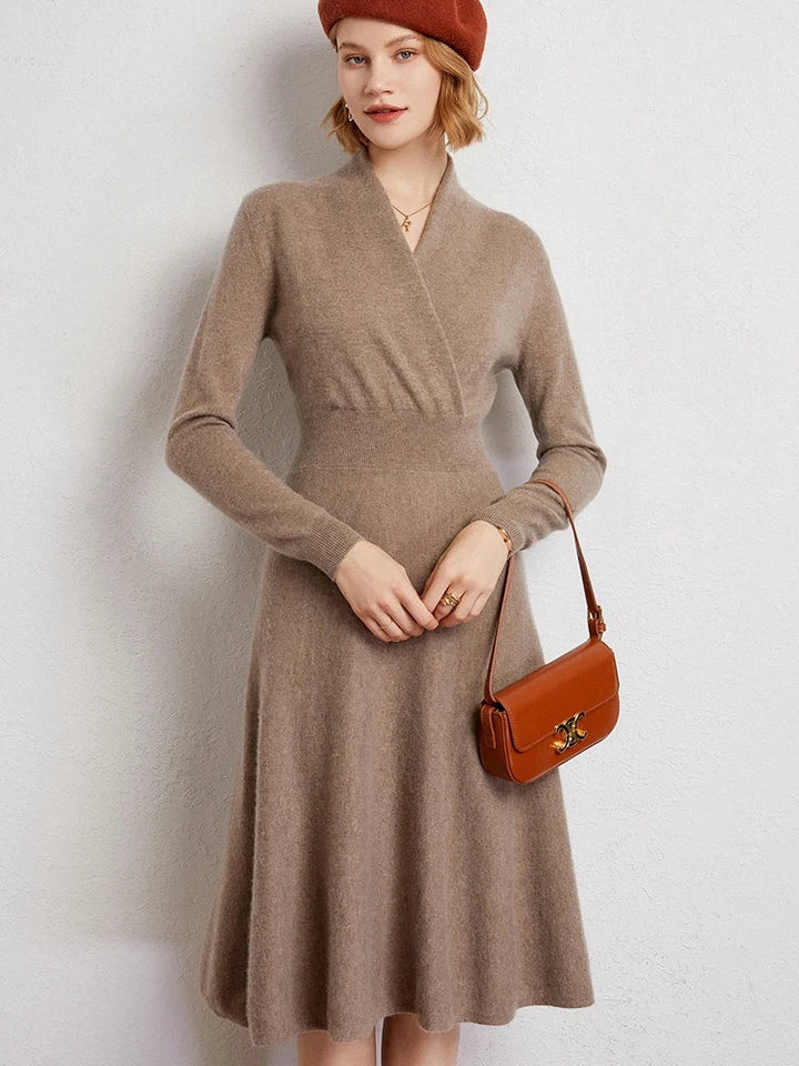 A woman wearing a light brown, knee-length cashmere dress with long sleeves, a matching beret, and a brown shoulder bag stands against a light-colored wall, smiling slightly.