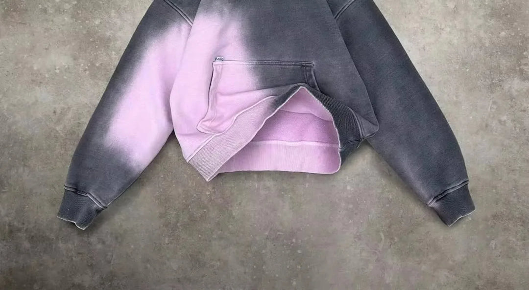 A cropped hoodie with a relaxed fit and front pocket, featuring a two-tone design in dark gray and light pink, laid flat on a textured gray surface.