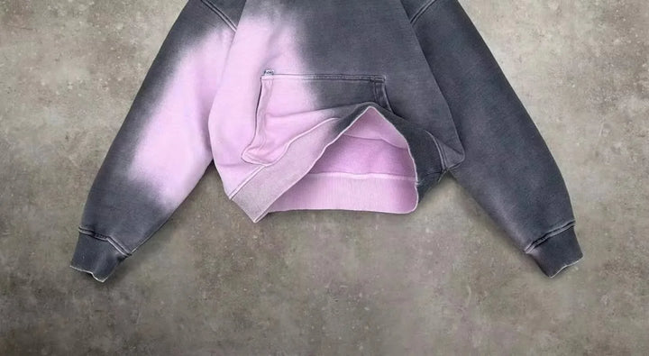 A cropped hoodie with a relaxed fit and front pocket, featuring a two-tone design in dark gray and light pink, laid flat on a textured gray surface.