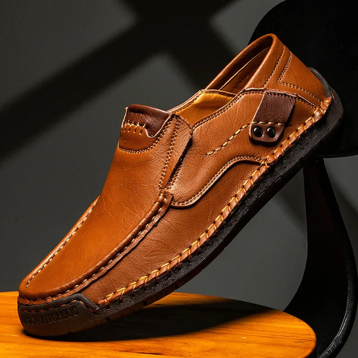 A stylish brown leather loafer with detailed stitching, textured sole, and decorative tab detail on the side, displayed against a dark background and resting on a wooden surface.