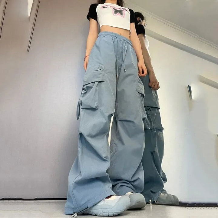A person stands indoors wearing light blue cargo pants with cargo pockets, gray chunky sneakers, and a cropped white and black t-shirt with a butterfly graphic. The low angle shot emphasizes the oversized pants.