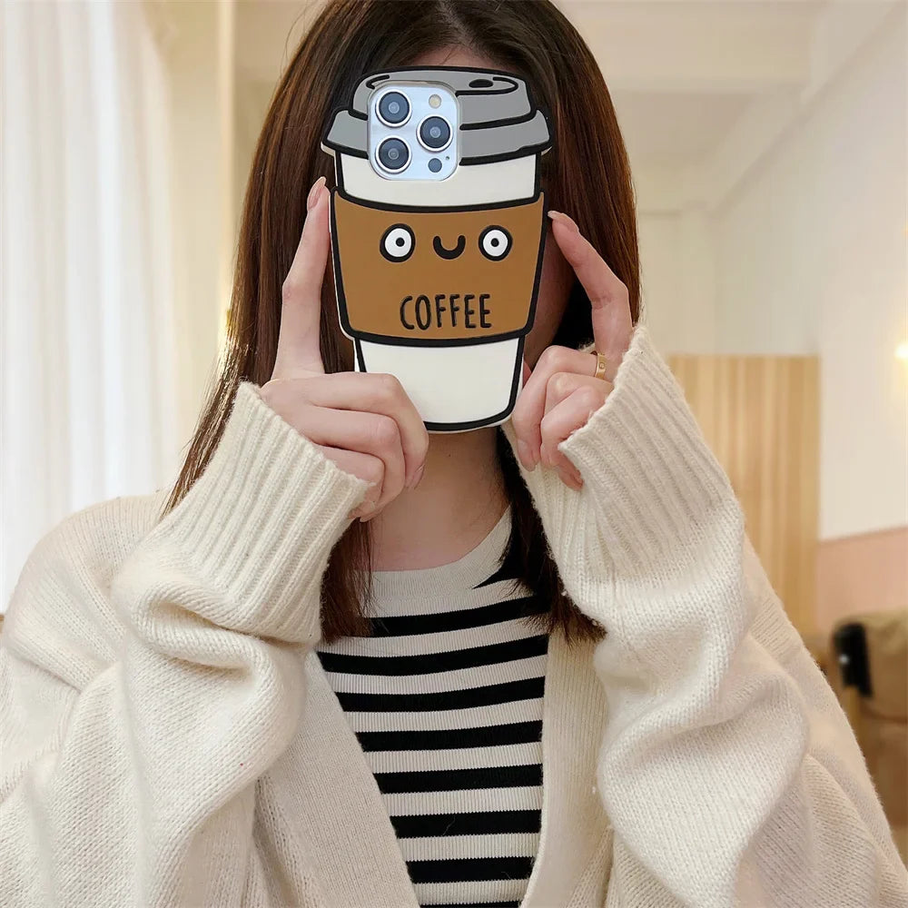 A person in a cream sweater holds a phone with a cute 3D coffee cup case in front of their face, covering it. The soft silicone case features a smiling face, the word COFFEE, and offers shockproof protection for your phone.