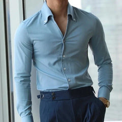 A person is standing by a window wearing a light blue slim fit shirt tucked into high-waisted navy blue trousers, accessorized with a gold wristwatch. The person’s face is not visible.
